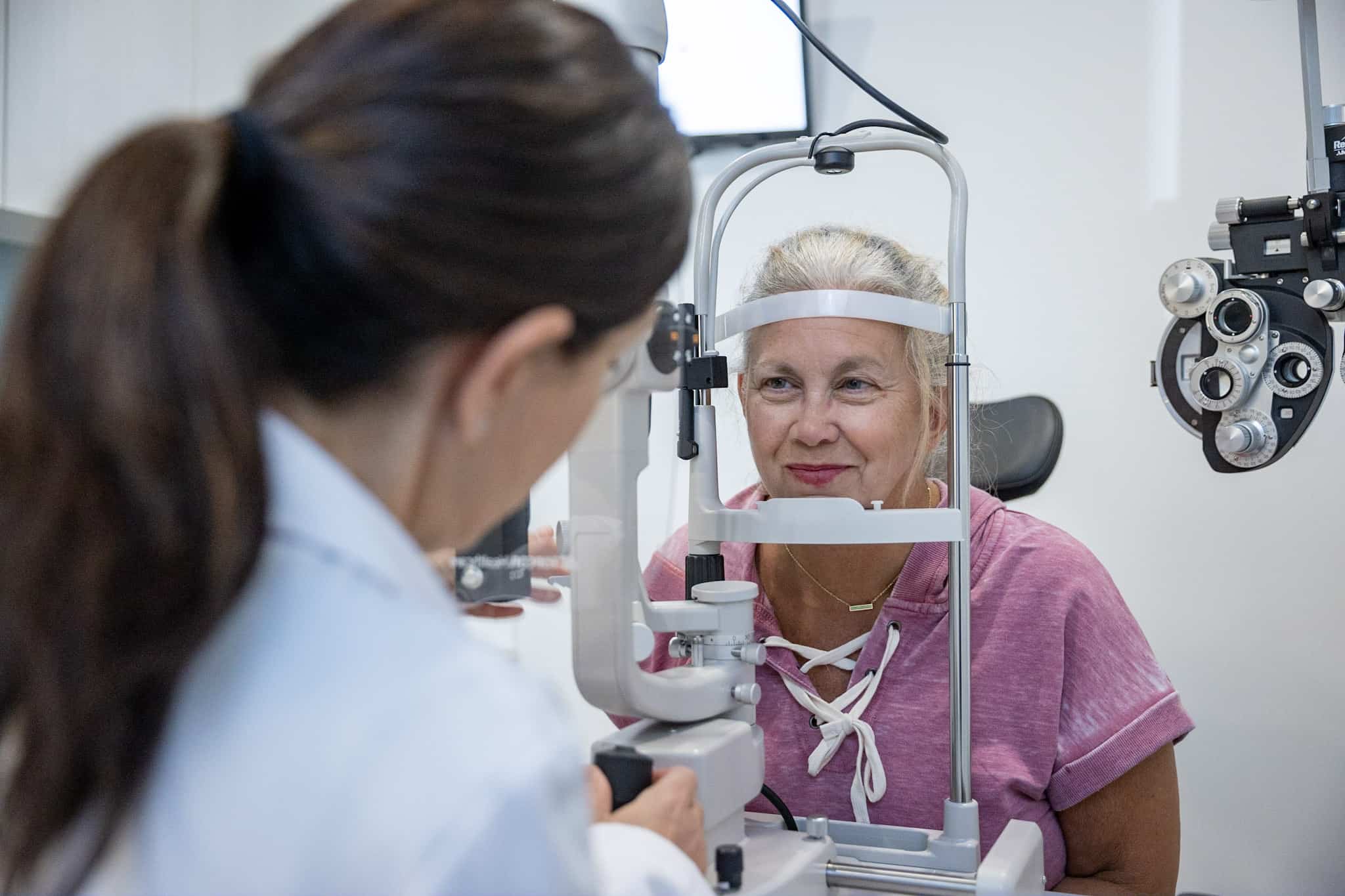 Dr. Helen J. Zervas behind positioning a slit lamp to examine a female senior patient’s eye health.