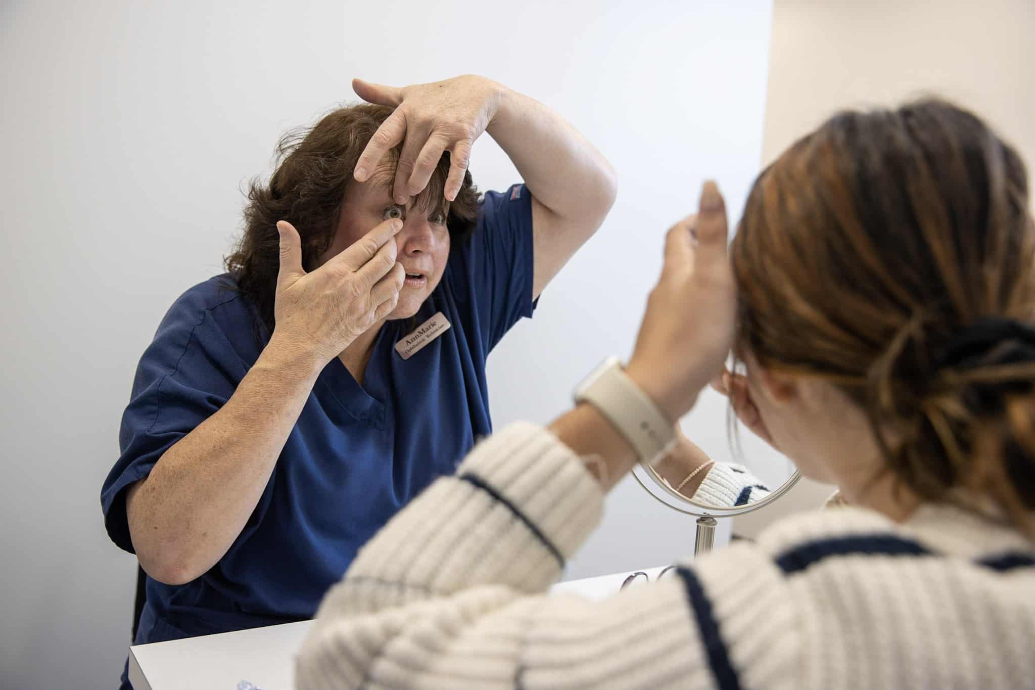 Optometric Technician AnnMarie using her hands to widen her eye to show to a female patient how to properly put in contacts.