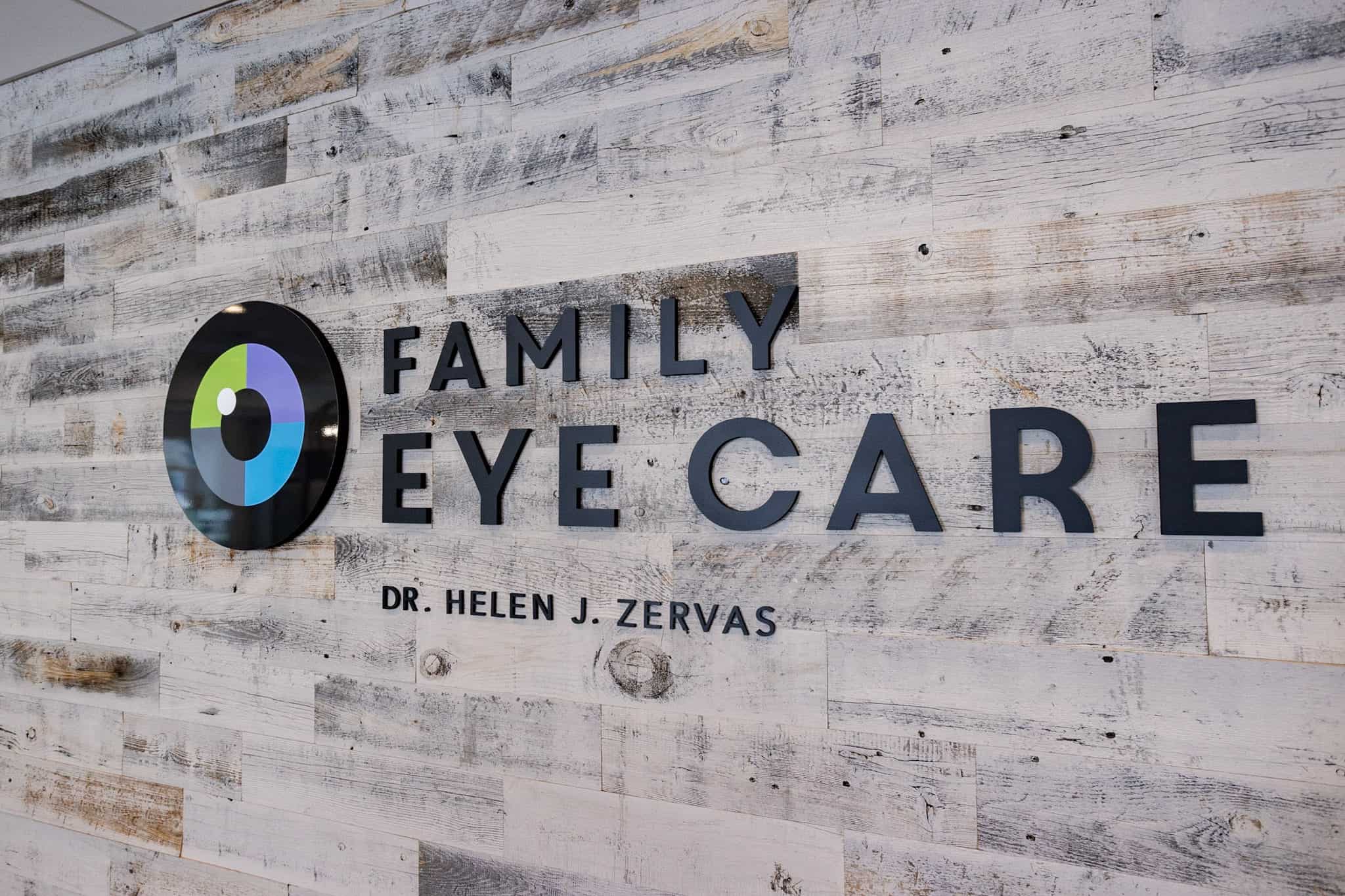 Gray wood grain wall with Family Eye Care logo followed by optometrist Dr. Helen J. Zervas’ name written underneath. 